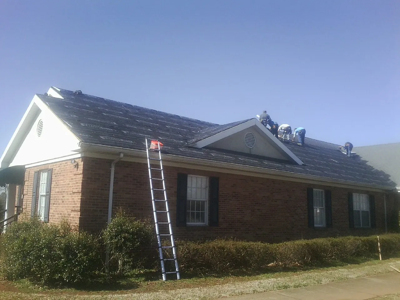 Crew tearing shingles off a brick ranch roof with a ladder set — Emergency Roof Repair work by our crew in Deltaville