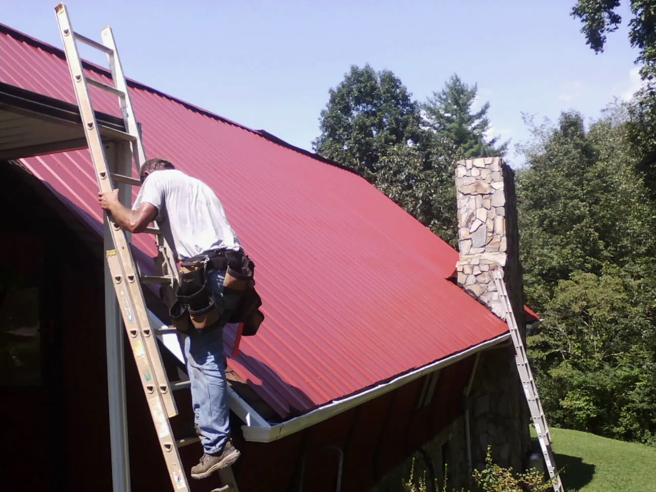 Roofer on a ladder beside a red metal roof and stone chimney — Soffit & Fascia Repair by Roofing Brothers in Deltaville